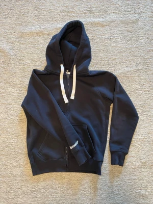 Mörkblå zip hoodie med vita snören från Rushemerch - Snygg mörkblå hoodie från rushemerch med dragkedja och vita snören i huvan. Klassisk modell med kängurufickor framtill och ribbade muddar vid ärmar och nederkant. Perfekt för en avslappnad och stilren look.