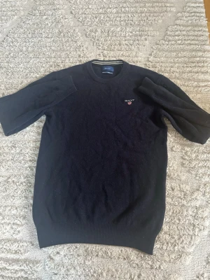 Svart stickad tröja från Gant - Klassisk mörkblå stickad tröja från Gant med rund halsringning och diskret logga broderad på bröstet. Tröjan har långa ärmar och ribbade muddar vid ärmslut och nederkant. Perfekt för en stilren och enkel look.