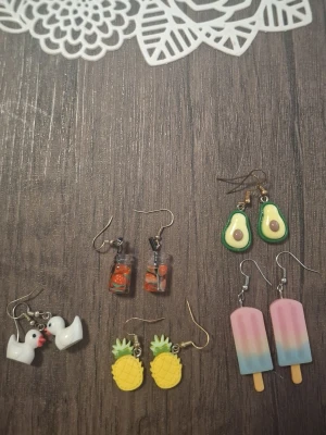Fem par färgglada örhängen - Säljer ett set med fem par örhängen i olika former: vita ankor, ananaser, avokadohalvor, glasspinnar i pastellfärger och små glas med apelsinskivor. Alla örhängen har krokar i metall och är tillverkade i plast med lekfull design. Perfekta för dig som gillar unika och roliga accessoarer.