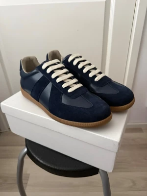 Maison Margiela mörkblå 43 - Snygga marinblå sneakers med beige sula och vita snören. Skorna har en mix av mocka och slätt läder, samt beige innerfoder. Klassisk låg modell som passar till det mesta och ger en clean look.
