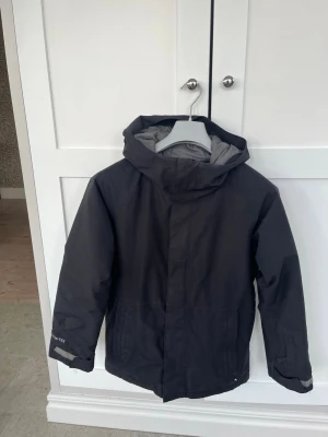 Svart gore tex vinterjacka från Burton - Snygg svart vinterjacka från Burton med huva och dragkedja. Jackan har GORE-TEX-tyg för extra väderskydd och en grå insida. Perfekt för blåsiga dagar,skidåkning och outdoor-aktiviteter. Enkel, stilren design med diskret logga på ärmen och ryggen.