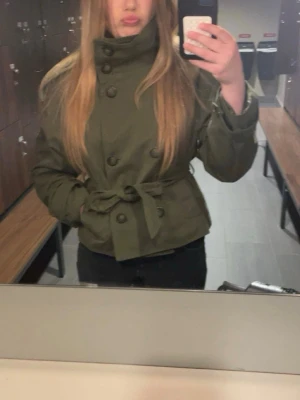 Olivgrön kort trenchcoat med knytskärp - Snygg olivgrön trenchcoat med dubbelknäppning och brett knytskärp i midjan. Jackan har en hög krage och stora knappar framtill. Kortare modell som ger en trendig siluett. Perfekt för dig som vill ha en stilren och cool look.