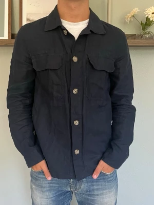 Mango Man Linneskjorta – 100% Linne | Marinblå - Storlek: S (EUR S / 180/96A) Passform: Regular — avslappnad utan att vara baggy Färg: Marinblå Material: 100% linne  En skjortjacka i 100% linne från Mango Man — marinblå med camp collar och två bröstfickor med lock. Linnet ger ett naturligt, luftigt fall som andas i varmare väder samtidigt som den funkar som ett skönt lager när kvällarna blir svalare.  Passar perfekt till t-shirt och jeans eller helt öppen över en enkel bas.  Modellen på bilden är 175 cm och väger 65 kg.