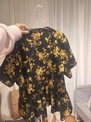 Svart blus med gult blommönster - Snygg svart blus med gult blommönster och volang nedtill. Blusen har korta ärmar och knytband i midjan för en feminin siluett. Perfekt för dig som vill ha en trendig och unik look med fina detaljer.