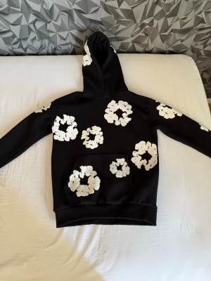 Svart hoodie från Denim Tears - Säljer en svart hoodie med stora vita blommönster över hela tröjan, inklusive huvan och ärmarna. Hoodien har en klassisk känguruficka framtill och ribbade muddar vid ärmslut och nederkant. Perfekt för dig som gillar en unik och trendig stil. S men passar Xs