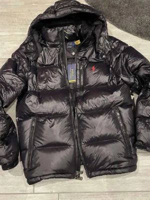 Svart pufferjacka från Polo Ralph Lauren - Säljer en svart pufferjacka från Polo Ralph Lauren storlek S men passar även M med röd logga på bröstet. Jackan har huva, dragkedja framtill och två sidofickor med dragkedja. Perfekt för kalla regniga och blåsiga dagar, nypris är runt 6000, kvitto kommer med