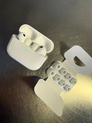 AirPods Pro 3  - Säljer ett par vita trådlösa in-ear hörlurar med laddningsetui och flera utbytbara silikonpluggar i olika storlekar. Hörlurarna har en stilren och kompakt design i blank plast och passar perfekt för musik, poddar och samtal på språng.