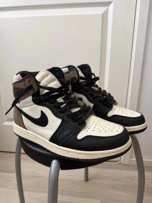 Air jordan 1 Mocha high  - Säljer ett par Nike Air Jordan 1 High sneakers i svart, vitt och brunt läder. Klassisk siluett med högt skaft, svarta snören och ikonisk Swoosh på sidan. Perforerad tåbox och vadderad plös för extra komfort.