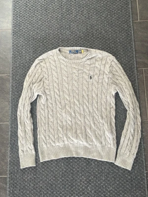 Ljusgrå kabelstickad tröja Polo Ralph Lauren - Kabelstickad tröja i ljusgrått från Polo Ralph Lauren. Klassisk rund halsringning, ribbade muddar och nederkant samt den ikoniska lilla loggan broderad på bröstet. Perfekt för dig som gillar stilrena och tidlösa plagg.