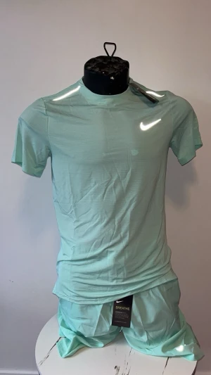 Mintgrönt träningsset från Nike - Snyggt mintgrönt träningsset från Nike med t-shirt och shorts. Setet har en enkel och sportig design med rund halsringning och korta ärmar. Tillverkat i lätt och ventilerande material, perfekt för träning eller gym. Reflekterande Nike-logga på bröstet.