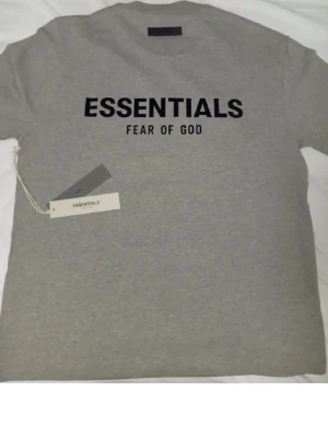 Grå Essentials Fear of God t-shirt passar S - Snygg grå t-shirt från Essentials Fear of God med svart tryck på bröstet. Klassisk passform och rund halsringning. Perfekt till jeans eller shorts för en clean och trendig look. 