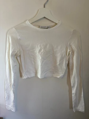 Vit croppad långärmad topp - Säljer en enkel och stilren vit croppad topp med långa ärmar och rund hals. Toppen är gjord i mjuk bomull och passar perfekt till jeans eller kjol för en clean look.