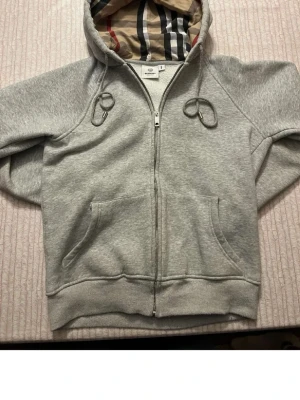 Grå zip hoodie med huva burberry - En klassisk grå zip hoodie med huva och fickor framtill. Tillverkad i mjukt sweatshirtmaterial som är perfekt för chill dagar. Ribbstickade muddar vid ärmslut och nederkant. Enkel och stilren design som passar till det mesta. Dragkedjan är svår stängd liten tyg bit som sitter där man stänger den. snörena är lite slitna blev så när jag tvätta tröjan annars är den som ny