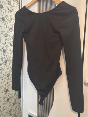 Svart långärmad body med djup rygg - Snygg svart body med långa ärmar och djup urringning i ryggen. Tillverkad i stretchigt material som sitter tight mot kroppen. Perfekt att styla med jeans eller kjol för en clean look.