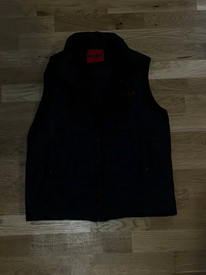 Svart dunväst från Hugo - Stilren svart dunväst från Hugo med hög krage och dragkedja framtill. Västen har diskret quiltad design och två sidofickor. Perfekt för lager-på-lager och ger en clean look till din outfit.