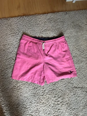 Rosa badbyxor från Polo Ralph Lauren - Säljer ett par snygga rosa badbyxor från Polo Ralph Lauren. De har elastisk midja med vit dragsko, sidofickor och en bakficka. Klassisk passform och broderad logga på vänster ben. Perfekta för stranden eller poolen.