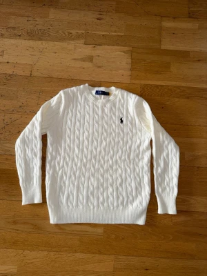 Vit stickad tröja Polo Ralph Lauren - Stickad vit tröja från Polo Ralph Lauren med rund halsringning och svart broderad logga på bröstet. Tröjan har långa ärmar och ribbade muddar vid ärmslut och nederkant. Perfekt för en klassisk och stilren look.
