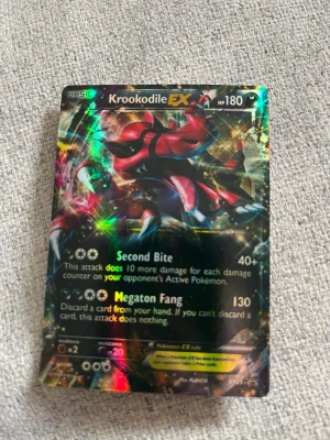 Pokémonkort Krookodile EX holografiskt - Säljer ett holografiskt Pokémonkort med Krookodile EX, 180 HP. Kortet har en glittrig yta och färger i svart, rött och gult. Det är tillverkat i laminerad papp och har attacker som Second Bite och Megaton Fang. Perfekt för dig som samlar eller spelar Pokémonkort.