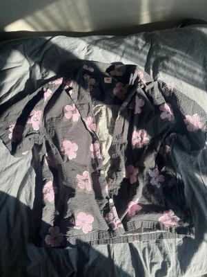 Svart skjorta med rosa blommor - Kortärmad skjorta i svart med stora rosa blommor över hela plagget. Skjortan har knappar framtill och en avslappnad passform. Materialet ser ut att vara bomull med en lätt strukturerad yta, perfekt för varmare dagar.