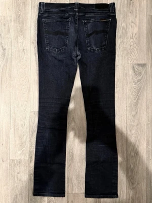 Nudie Jeans Tube Tom Mörkgrå/Svarta W32/L32 - Slim Fit - Nudie Jeans Tube Tom ”Org. Back In Black” mörkgrå/svarta jeans med slim fit-passform, normalhög midja och avsmalnande ben, storlek W32/L32, midja 40 cm, ytterbenslängd 99,5 cm, benöppning 16,5 cm, modellen är 185cm & väger 65kg, i fint skick.