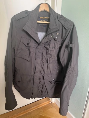 Mörkgrå vindjacka från Woolrich - Snygg mörkgrå vindjacka från Woolrich med flera praktiska fickor framtill, hög krage och axelklaffar. Jackan har dragkedja och tryckknappar, samt detaljerade sömmar på ärmarna. Tillverkad i ett lätt och slitstarkt material, perfekt för blåsiga dagar.Perfelt ha över en hoodie och ännu bättre nu till hösten