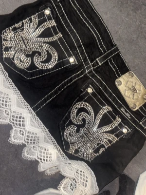 Svart jeanskjol med spets och strass - Unik kjol med stora bakfickor dekorerade med glittrande strassmönster och vita kontrastsömmar. Nederkanten pryds av bred vit spets som ger en extra detalj. Kjolens metallmärke och nitar ger en cool touch.