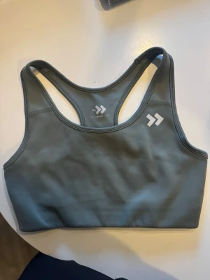Grå sport-bh med brottarrygg - Snygg grå sport-bh med brottarrygg och minimalistisk design. Tillverkad i ett stretchigt och mjukt material som passar perfekt för träning. Har en liten vit logga framtill och breda axelband för extra stöd.