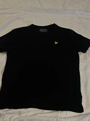 Svart t-shirt från Lyle & Scott - Klassisk svart t-shirt från Lyle & Scott med gul logga på bröstet. T-shirten har rund hals och korta ärmar. Perfekt basplagg i mjuk bomull som funkar till det mesta.