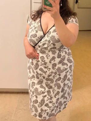 Vintage omlottklänning med paisleymönster - Snygg vit omlottklänning med brunt paisleymönster och knyt i midjan. Klänningen har korta ärmar och v-ringning, perfekt för en trendig och avslappnad look. Tillverkad i mjukt bomullsmaterial som känns skönt mot huden.