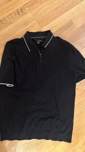 Svart pike med zip från M&S - Svart pike från M&S i bomullsmix med vit piping på krage och ärmslut. Halvlång dragkedja framtill och korta ärmar ger en clean och sportig vibe. Perfekt till jeans eller kjol för en enkel men snygg look.