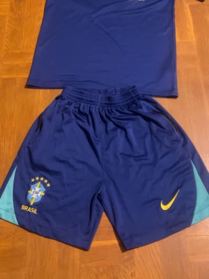 Blått Brasilien träningsset från Nike - Säljer ett blått träningsset från Nike med Brasilien-logga och Dri-Fit material. Setet består av en t-shirt och shorts med gula och turkosa detaljer samt Nike Swoosh. Perfekt för fotbollsträning eller gymmet.