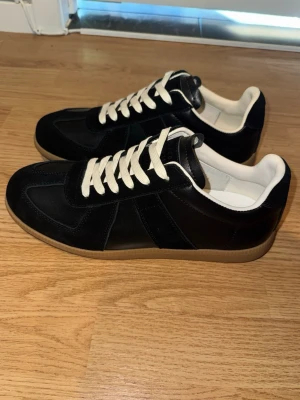 Svarta Maison Margiela gats  - Stilrena svarta sneakers med ovandel i mocka och läder, vita snören och kontrasterande ljusbrun gummisula. Klassisk low-top modell med vit insida och diskret design som passar till det mesta. Perfekta för dig som gillar en clean och tidlös look.