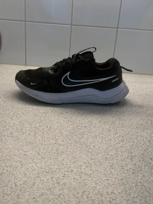Svarta Nike löparskor med vit sula - Snygga svarta Nike sneakers med vit sula och klassisk vit swoosh på sidan. Ovandelen är i mesh och syntet för bra andningsförmåga och komfort. Lågt skaft och snörning för en sportig look. Perfekta för löpning eller träning.