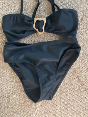 Svart bikini med gulddetalj - Stilren svart bikini med bandeau-topp och snygg guldfärgad detalj framtill. Inlägg i bh:n. Klassisk passform på både överdel och underdel, perfekt för strand eller pool. Tillverkad i ett mjukt och stretchigt syntetmaterial som sitter bekvämt hela dagen. Aldrig använd, endast provad.
