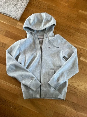 Grå hoodie med dragkedja Tommy Hilfiger - Snygg grå hoodie från Tommy Hilfiger med dragkedja framtill och liten logga på bröstet. Tillverkad i mjukt sweatshirtmaterial, har huva med dragsko och två fickor fram. Perfekt för en avslappnad och stilren look.