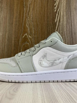 Nike Air Jordan 1 Low grå/vit - Snygga Air Jordan 1 Low sneakers i ljusgrått och vitt med camo-detaljer på swoosh-loggan. Skorna har en klassisk låg siluett, ovandel i läder och textil samt broderad Air Jordan-logga på hälen. Perfekta för dig som gillar stilrena och trendiga sneakers. Kostar 3000+ där ute 