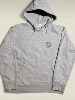 Grå hoodie från Nudie Jeans - Klassisk grå hoodie från Nudie Jeans med liten logga i rött på bröstet. Hoodien har huva med dragsko, magficka och ribbade muddar vid ärmar och nederkant. Tillverkad i mjuk bomullsblandning, perfekt för en avslappnad stil. Hör av er vid frågor. 
