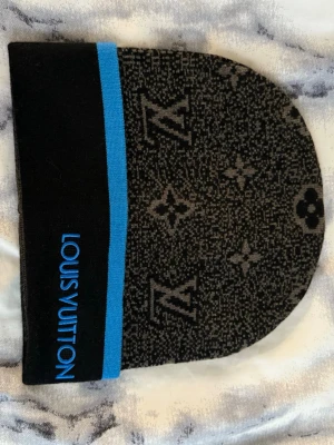 Svart mössa - Snygg svart och grå mössa med klassiskt monogrammönster och en blå rand vid kanten. Märkets logga är broderad i blått på sidan. Mössan är stickad och har en modern, avslappnad passform.