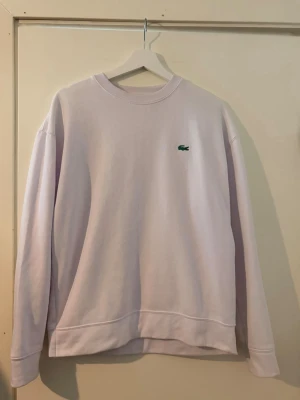 Lacoste sweatshirt - En stilren vit sweatshirt från Lacoste med klassisk rund halsringning och det ikoniska gröna krokodilmärket broderat på bröstet. Tröjan har långa ärmar och ribbade muddar vid ärmslut och nederkant. Perfekt för en avslappnad look.