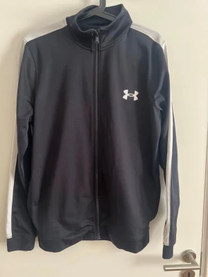 Svart tracksuit från Under Armour - Säljer ett svart tracksuit från Under Armour med vita detaljer längs ärmar och ben. Setet består av en zip-jacka med hög krage och matchande byxor med dragsko i midjan. Båda plaggen har Under Armour-logga på bröst och lår. Perfekt för gymmet eller löprundan.