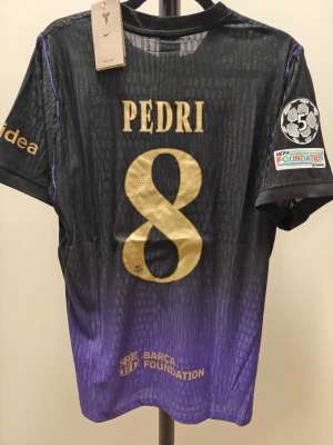 Barça  spelar version borta 26/27 Pedri #8 svart/lila - Snygg FC Barcelona matchtröja i svart och lila med guldiga detaljer. Pedri #8 på ryggen, Champions League-märke på ärmen och Barca Foundation-logga. Tröjan har klubbmärke och Spotify-logga framtill. Perfekt för dig som älskar fotboll och vill sticka ut på planen.