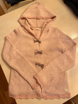 Ljusrosa stickad hoodie med rosetter - Supersöt ljusrosa stickad hoodie från Odd Molly med diskret mönster, tre dekorativa rosetter framtill och broderad logga på ärmen. Tröjan har huva och fina detaljer längs kanterna. Perfekt för dig som gillar en gullig och avslappnad stil.