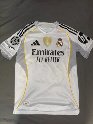 Real Madrid matchtröja Adidas vit - Säljer en vit Real Madrid matchtröja från Adidas med svarta detaljer och gula linjer. Tröjan har klubbmärke, Adidas-logga och sponsortryck på bröstet samt FIFA-märke. Korta ärmar med svarta ränder och patch på ena ärmen. Perfekt för fotbollsfans!