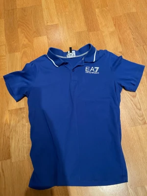 Blå pikétröja från EA7 Emporio Armani - Snygg blå pikétröja från EA7 Emporio Armani med vit logga på bröstet och vit kant på kragen. Klassisk pikédesign med korta ärmar och krage. Perfekt till en avslappnad men ändå stilren look. Storleken hade inte passat en 14 åring tycker jag utan den är lite mindre än vad lappen säger.