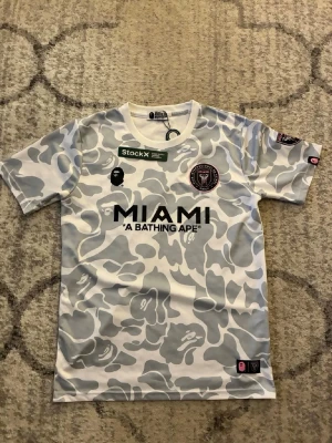 Inter Miami x BAPE camo t-shirt - Säljer en limited edition Inter Miami t-shirt från A Bathing Ape med gråvitt camomönster. Stor MIAMI-tryck på bröstet, BAPE-logga och Inter Miami CF-emblem. Perfekt för dig som gillar streetwear och fotbollströjor med unik design.