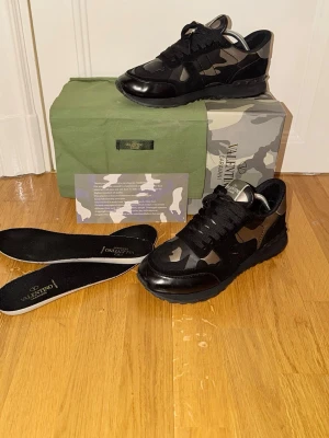 Valentino camo sneakers i svart - Snygga sneakers från Valentino i svart med mörkgrått camo-mönster. Skorna har svarta snören, detaljer i läder och textil samt en robust sula. Perfekta för dig som vill ha en unik och trendig look. Kommer med originalkartong och extra innersulor.