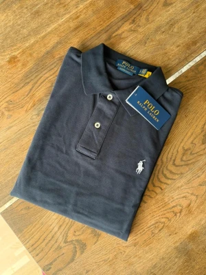 Mörkblå pikétröja från Polo Ralph Lauren - Snygg mörkblå pikétröja från Polo Ralph Lauren med klassisk krage och två knappar framtill. Tröjan har korta ärmar och den ikoniska broderade loggan i vitt på bröstet. Perfekt för en stilren och avslappnad look.