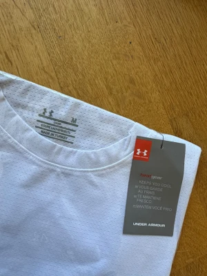 Vit tränings-tshirt från Under Armour - Säljer en vit kortärmad t-shirt från Under Armour med diskret logga på bröstet. Tillverkad i ett lätt och ventilerande material som passar perfekt för träning. Klassisk rund hals och sportig look.