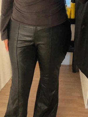 Svarta utsvängda skinnbyxor - Ett par svarta skinnbyxor med bootcut och hög midja. Byxorna har en söm framtill som ger en extra detalj. (Byxorna har en dragkedja vid sidan som är lite sönder men inget som syns) massor som M