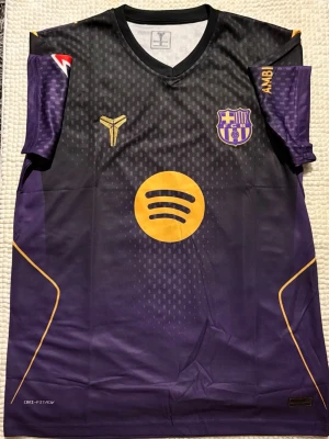 Lila FC Barcelona matchtröja - Snygg lila FC Barcelona matchtröja med guldfärgade detaljer och Spotify-logga på bröstet. Tröjan har klubbmärke på vänster sida och ett sportigt mönster över hela framsidan. Tillverkad i ett lätt och ventilerande funktionsmaterial. Passar L också. Köpt utom lands